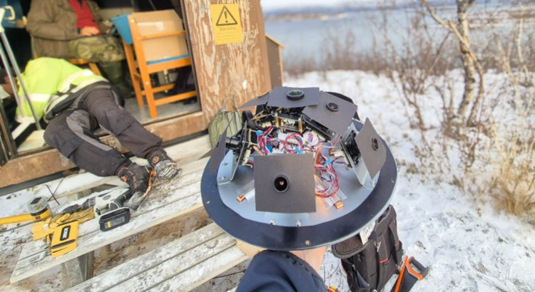 Meteorkamera utan sitt skyddande hölje. I bakgrunden mätstation i vintrigt Abisko med öppen dörr, mängder av verktyg och två av IRFs medarbetare i arbete. Meteorkamera utan sitt skyddande hölje. I bakgrunden mätstation i vintrigt Abisko med öppen dörr, mängder av verktyg och två av IRFs medarbetare i arbete.