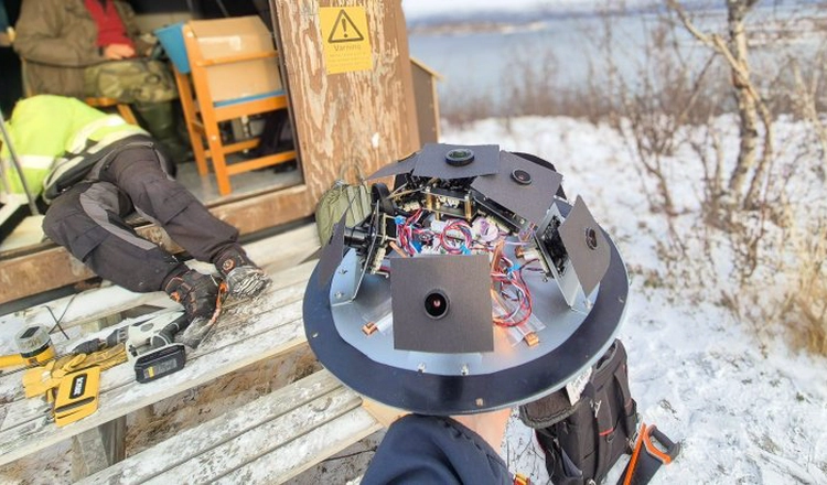 Meteorkamera utan sitt skyddande hölje. I bakgrunden mätstation i vintrigt Abisko med öppen dörr, mängder av verktyg och två av IRFs medarbetare i arbete. Meteorkamera utan sitt skyddande hölje. I bakgrunden mätstation i vintrigt Abisko med öppen dörr, mängder av verktyg och två av IRFs medarbetare i arbete.