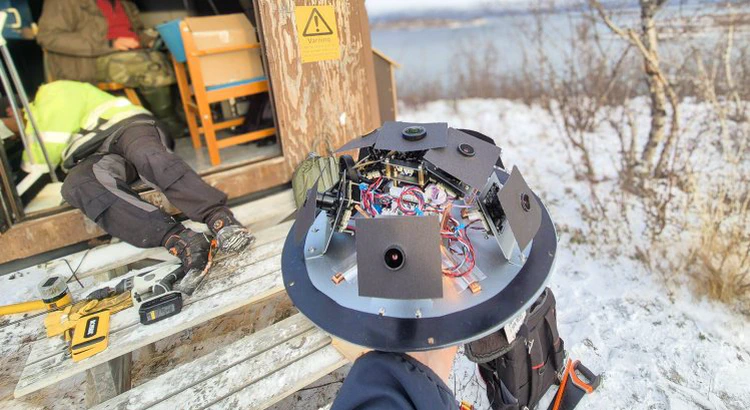 Meteorkamera utan sitt skyddande hölje. I bakgrunden mätstation i vintrigt Abisko med öppen dörr, mängder av verktyg och två av IRFs medarbetare i arbete. Meteorkamera utan sitt skyddande hölje. I bakgrunden mätstation i vintrigt Abisko med öppen dörr, mängder av verktyg och två av IRFs medarbetare i arbete.